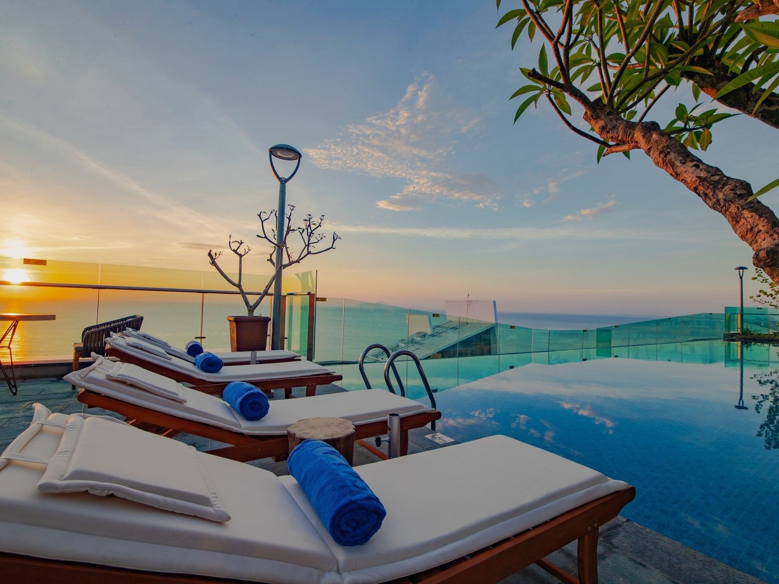 Rooftop pool deck at sunset with white loungers, palm fronds and a glass edge over the city