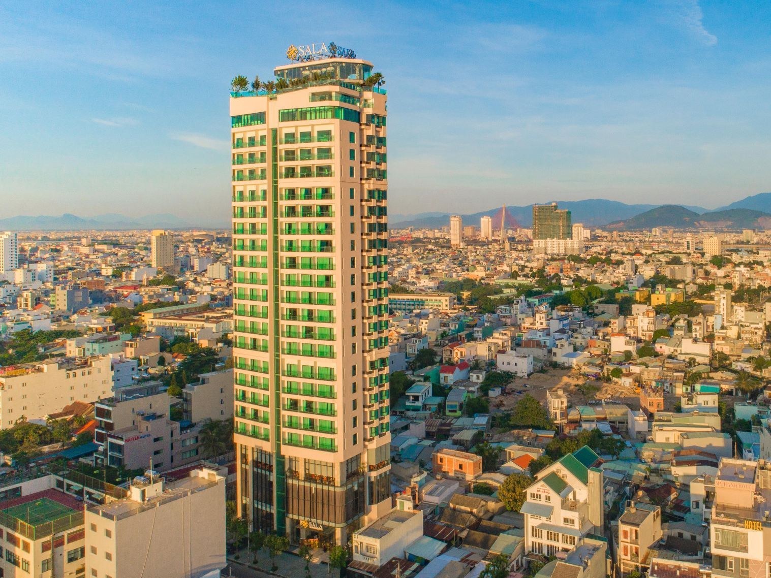 Slim hotel tower with green-tinted windows rising above low city rooftops, with mountains on the horizon