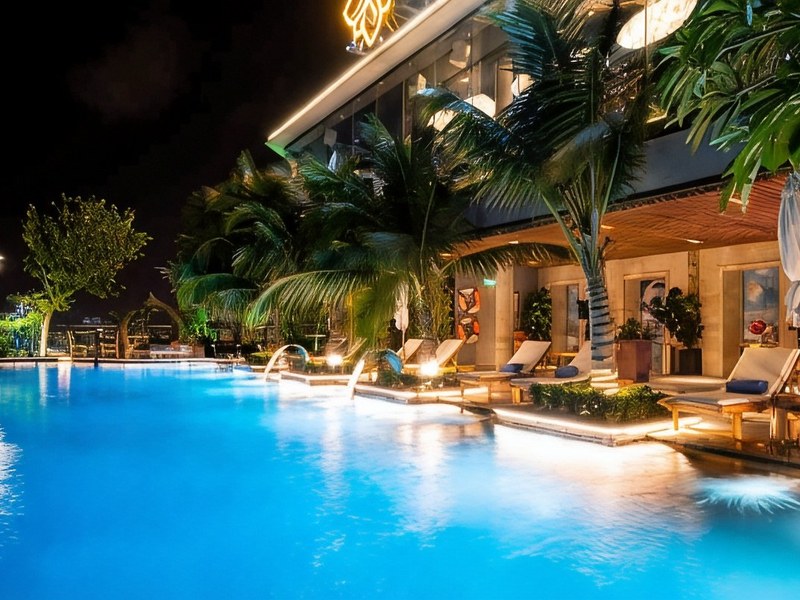 Rooftop infinity pool lit up at night, palms and loungers along a glass edge