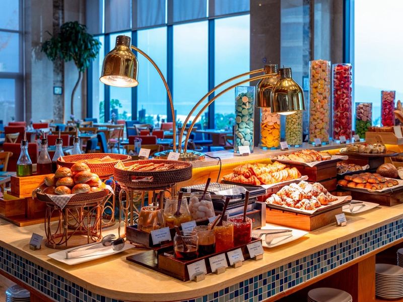 Breakfast buffet counter with hot dishes under brass lamps beside tall windows